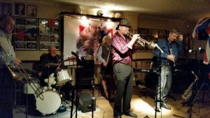 'Hershey and the Hotshots' playing for the first time at Farnborough Jazz Club on 4th December 2015. (LtoR) Paul Taylor (trombone), (Keith Grant has sit-in on drums), Roger Curphey (double bass), Peter Leonard (trumpet&vocs), Tim Huskisson (clarinet&alto sax) and (out of view) Andrew Clancy (piano) and drummer Graham Caulicott dances with Jill.  Photo by Peter Marr.